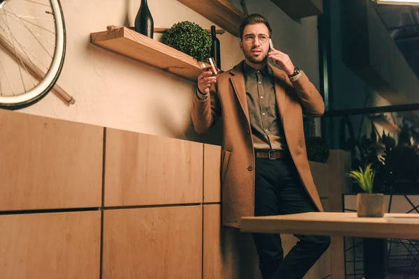 Portrait of stylish man in jacket with glass of whiskey talking on smartphone in cafe — Stock Photo