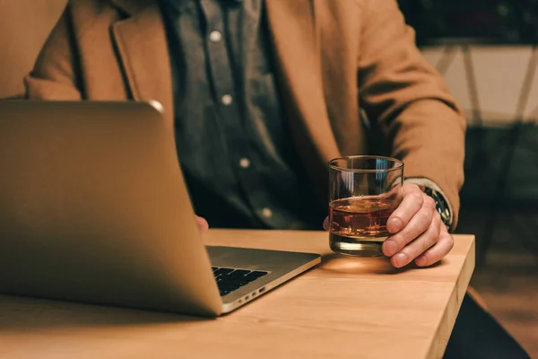 Tiro cortado de homem com copo de uísque sentado à mesa com laptop — Fotografia de Stock