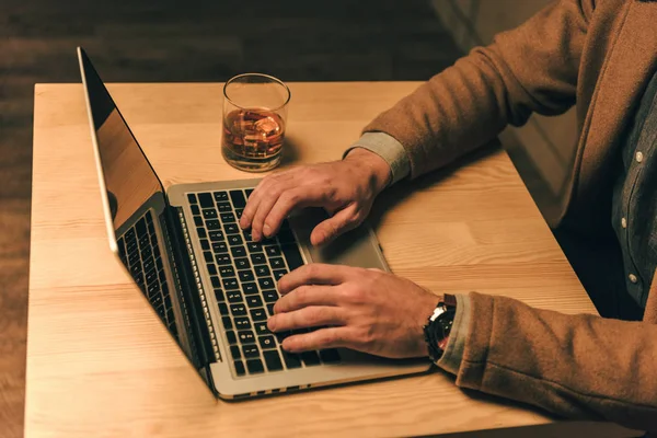 Cropped shot of man typing on laptop at table with glass of whiskey — Stock Photo