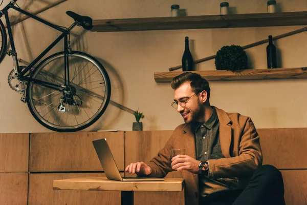 Ritratto di uomo sorridente con bicchiere di whisky utilizzando il computer portatile a tavola nel caffè — Foto stock