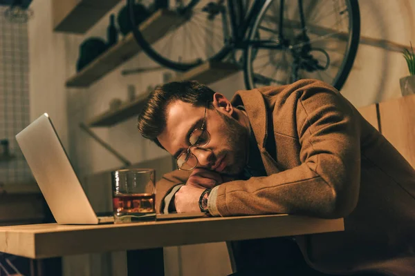 Уставший мужчина спит на столе со стаканом виски и ноутбуком в кафе — стоковое фото