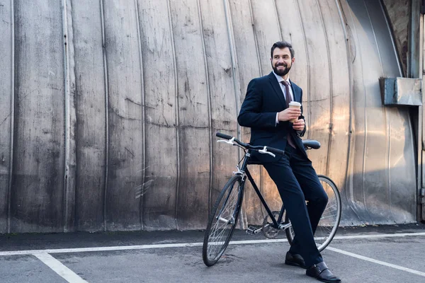 Homme d'affaires souriant avec tasse de café jetable appuyé sur le vélo dans la rue — Photo de stock