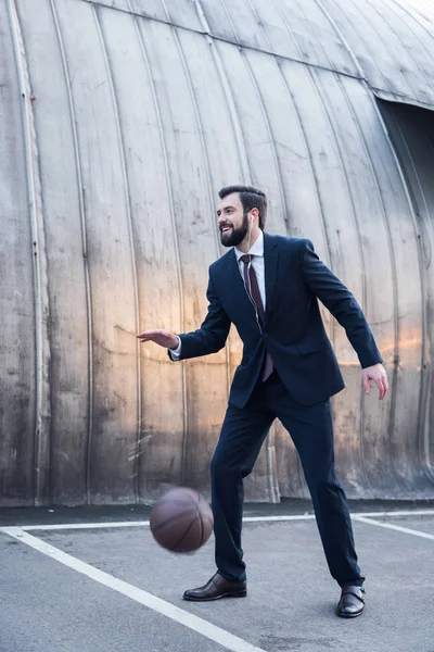 Side view of cheerful businessman in earphones playing basketball on street — Stock Photo