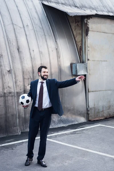 Joven hombre de negocios sonriente en traje apuntando mientras juega al fútbol en la calle - foto de stock