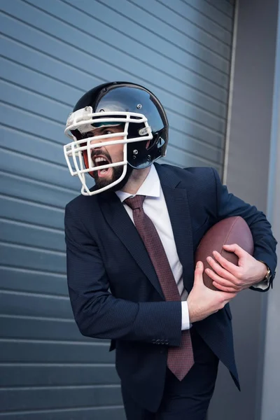 Joven hombre de negocios enojado en traje y casco de rugby con pelota en las manos en la calle - foto de stock