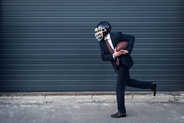 Side view of young businessman in suit and rugby helmet with ball in hands running on street — Stock Photo