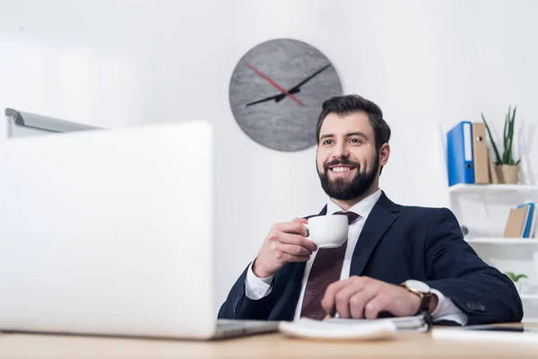 Foco seletivo de empresário alegre em terno beber café no local de trabalho no escritório — Fotografia de Stock