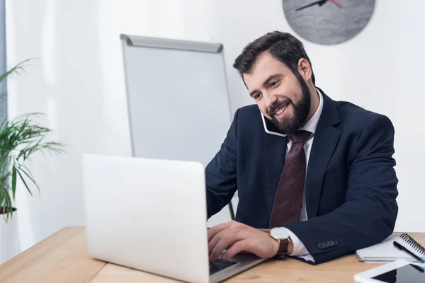 Retrato de homem de negócios sorridente falando no smartphone enquanto trabalhava no laptop no escritório — Fotografia de Stock