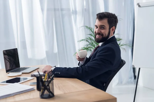 Cheerful businessman in suit drinking coffee at workplace with laptop in office — Stock Photo