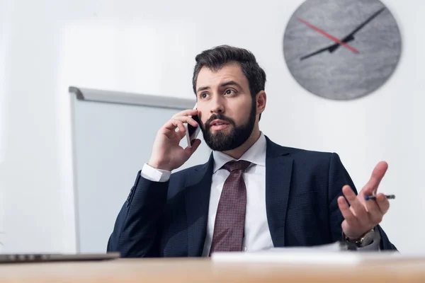 Portrait of young businessman talking on smartphone at workplace in office — Stock Photo