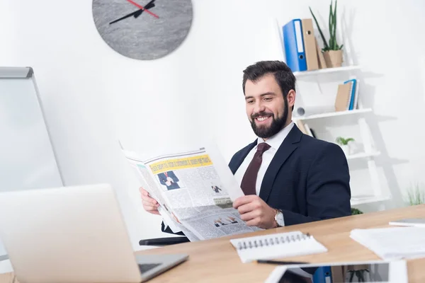 Porträt eines lächelnden Geschäftsmannes im Anzug, der am Arbeitsplatz im Büro Zeitung liest — Stockfoto