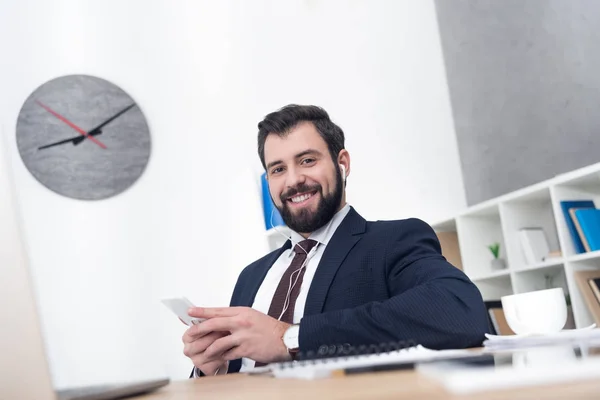 Businessman listening music in earphones with smartphone at workplace in office — Stock Photo