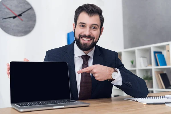 Retrato de empresario alegre apuntando a la computadora portátil en el lugar de trabajo en la oficina - foto de stock