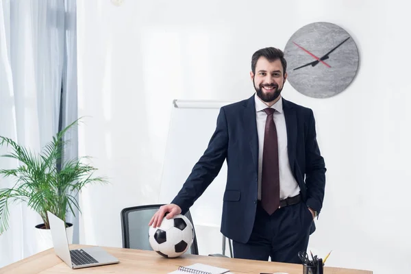 Homem de negócios sorridente em terno com bola de futebol no local de trabalho no escritório — Fotografia de Stock