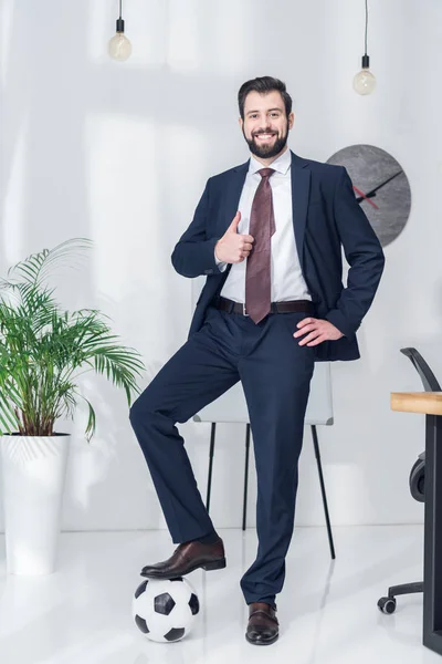 Alegre hombre de negocios de pie con una pierna en la pelota de fútbol y mostrando el pulgar en la oficina - foto de stock