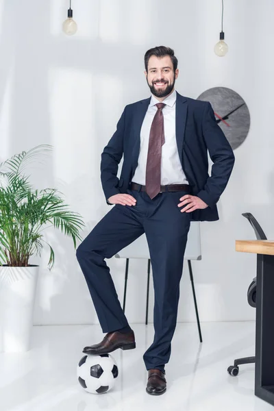 Homem de negócios alegre akimbo de pé com uma perna na bola de futebol no escritório — Fotografia de Stock