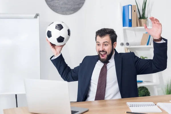 Aufgeregter Geschäftsmann im Anzug mit Fußballball blickt am Arbeitsplatz im Büro auf den Laptop-Bildschirm — Stockfoto