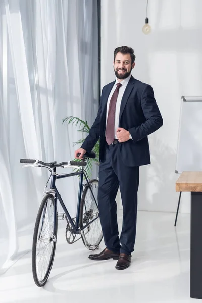 Smiling businessman in suit standing near bicycle in office — Stock Photo