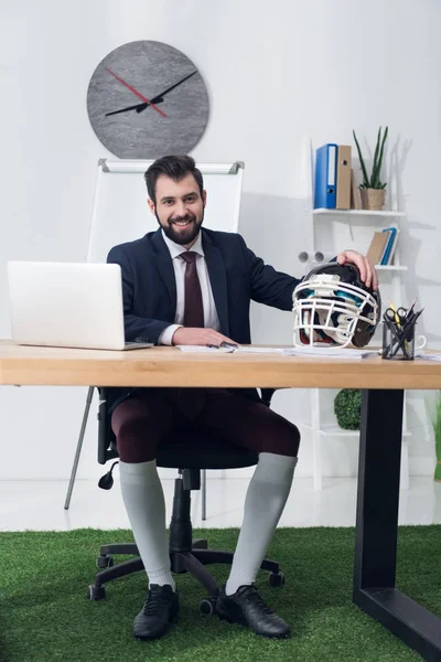 Joven empresario sentado en el lugar de trabajo con casco de rugby en la oficina - foto de stock