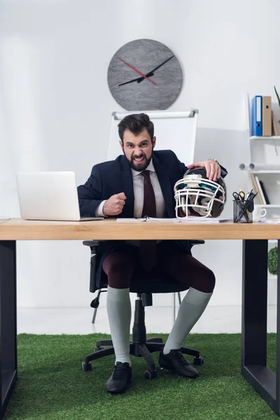 Angry businessman sitting at workplace with rugby helmet in office — Stock Photo