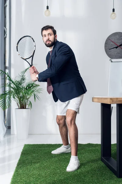 Young businessman in jacket and shorts playing tennis in office — Stock Photo