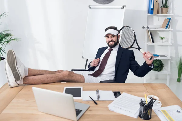 Jovem empresário em boné com raquete de tênis descansando no local de trabalho no escritório — Fotografia de Stock
