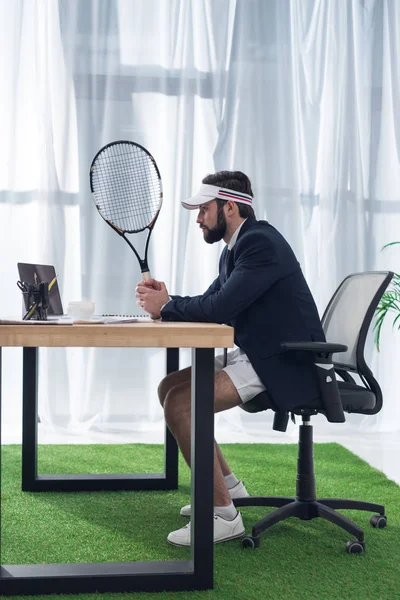 Vue latérale de l'homme d'affaires en casquette avec raquette de tennis sur le lieu de travail au bureau — Photo de stock