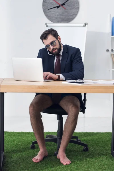 Businessman in jacket and shorts talking on smartphone at workplace — Stock Photo
