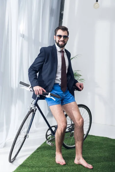 Smiling businessman in jacket and shorts leaning on bicycle in office — Stock Photo