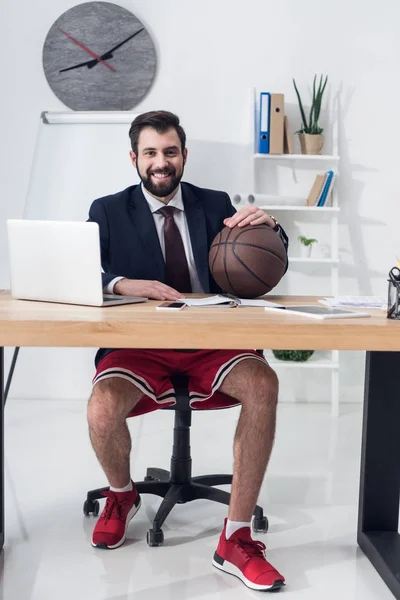Smiling businessman in shorts and jacket with basketball ball sitting at workplace — Stock Photo