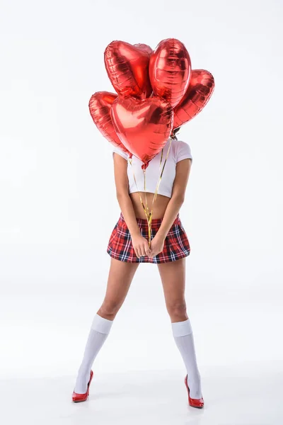 Sexy schoolgirl covering face with helium balloons in shape of hearts isolated on white — Stock Photo