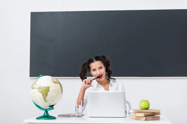 Young seductive teacher sitting at workplace with globe and laptop in classroom — Stock Photo