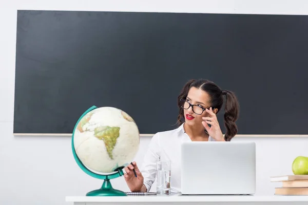 Вдумчивый сексуальный учитель, сидящий на рабочем месте с глобусом и ноутбуком в классе — стоковое фото