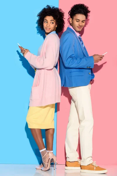 Joven pareja amerciana sonriente africana con teléfonos parados espalda con espalda sobre fondo rosa y azul - foto de stock
