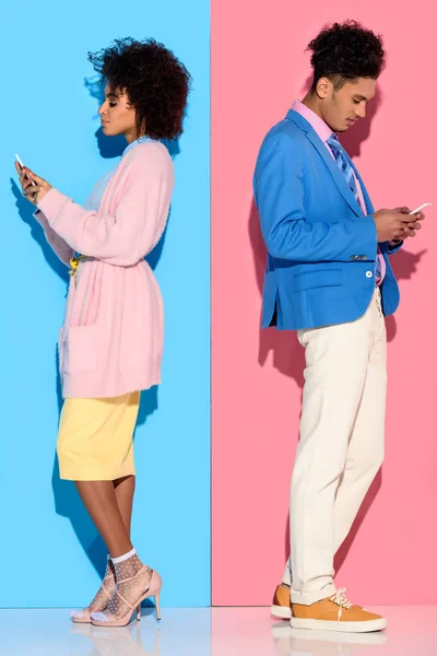 Joven pareja amerciana sonriente africana con teléfonos parados espalda con espalda sobre fondo rosa y azul - foto de stock