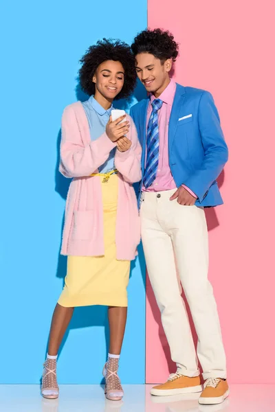 Jovem casal afro-americano olhando para a tela do telefone em fundo rosa e azul — Fotografia de Stock