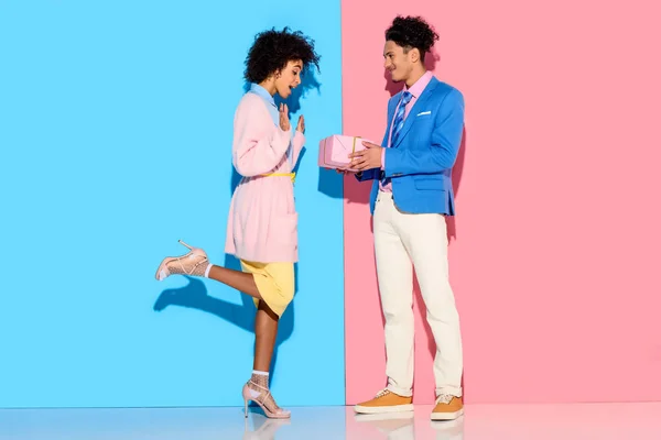 Junge afrikanisch-amerikanische Mann gibt Mädchen Geschenk-Box auf rosa und blauen Hintergrund — Stockfoto
