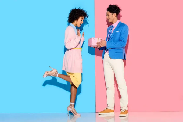 Jeune homme afro-américain donne boîte cadeau fille sur fond rose et bleu — Photo de stock