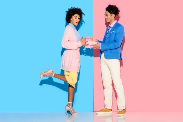 Jeune homme afro-américain donne boîte cadeau fille sur fond rose et bleu sur fond rose et bleu — Photo de stock