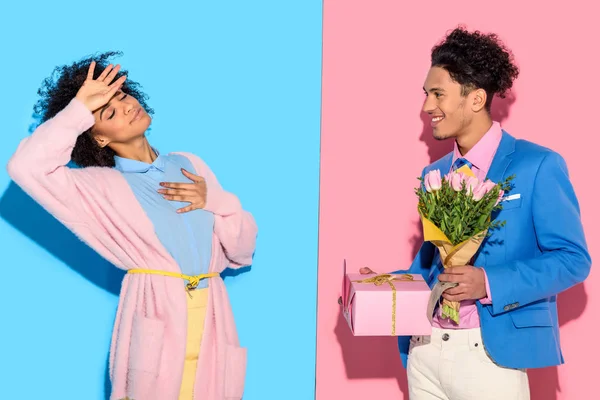 Arfican woman with hand on forehead while man presenting flowers on pink and blue background — Stock Photo