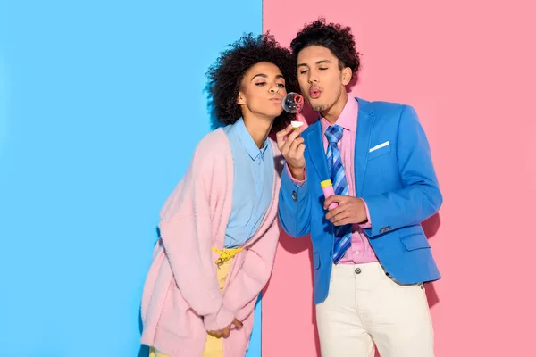 Afrikanisches Paar hat Spaß und bläst Seifenblasen auf rosa und blauem Hintergrund — Stockfoto