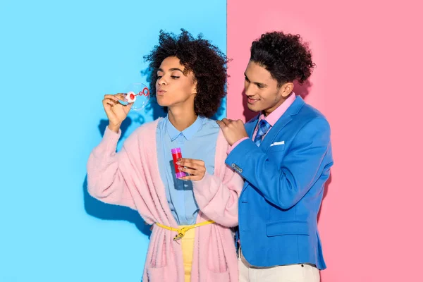 Casal africano se divertindo com bolhas de sabão no fundo rosa e azul — Fotografia de Stock