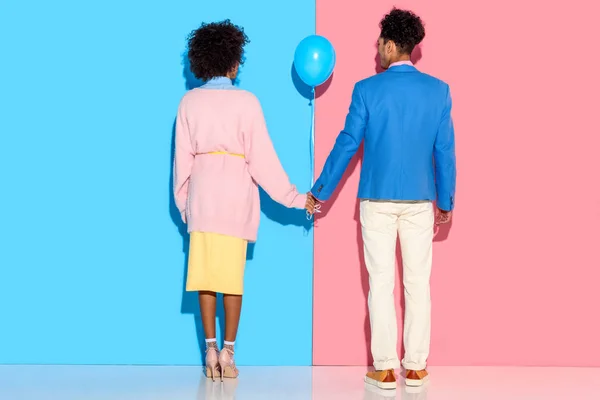 Vista trasera de pareja joven cogida de la mano con globo aerostático sobre fondo rosa y azul - foto de stock
