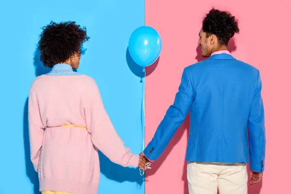 Vista trasera de pareja cogida de la mano con globo aerostático sobre fondo rosa y azul - foto de stock