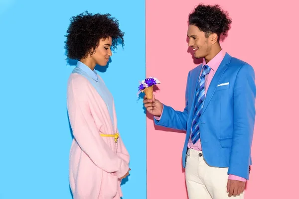 Bel homme présentant des fleurs à une jolie femme africaine sur fond rose et bleu — Photo de stock