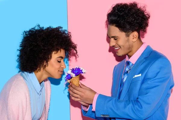 Schöner Mann überreicht Blumen an attraktive afrikanische Frau auf rosa und blauem Hintergrund — Stockfoto