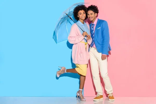 Couple souriant debout près de l'autre sous le parapluie et regardant la caméra sur fond rose et bleu — Photo de stock