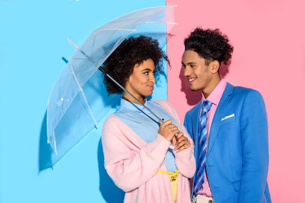 Pareja sonriente de pie cerca la una de la otra bajo paraguas sobre fondo rosa y azul - foto de stock