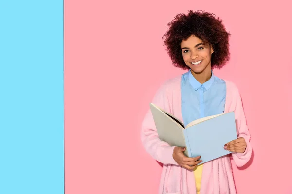 Portrait of smiling african american woman with book in hands looking at camera against pink and blue wall — Stock Photo