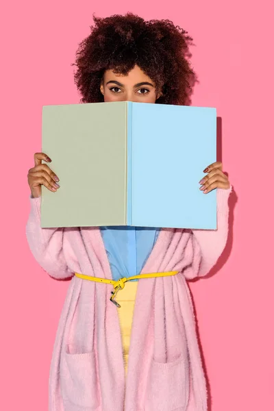 Obscured view of african american woman with book in hands looking at camera with pink wall backdrop — Stock Photo